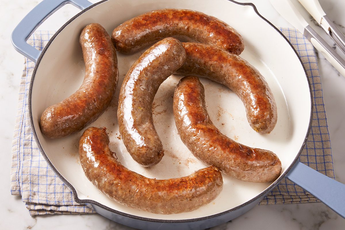 Six browned sausages are cooking in a white frying pan with light blue handles, placed on a checked cloth on a marble countertop. Metal tongs rest nearby.