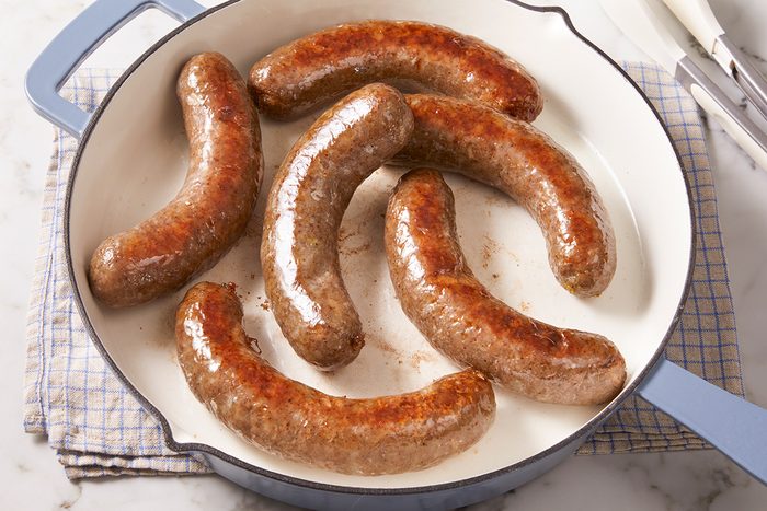 Six browned sausages are cooking in a white frying pan with light blue handles, placed on a checked cloth on a marble countertop. Metal tongs rest nearby.