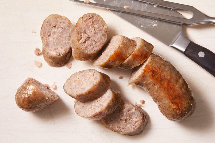 Sliced cooked sausage on a cutting board, with a knife and serving fork placed nearby. The sausage pieces are arranged in a casual manner.