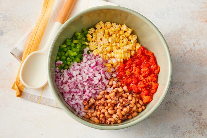 A bowl filled with chopped green bell pepper, corn, diced tomatoes, red onion, and black-eyed peas, arranged in sections. A white spoon and kitchen towel are beside the bowl on a light surface.