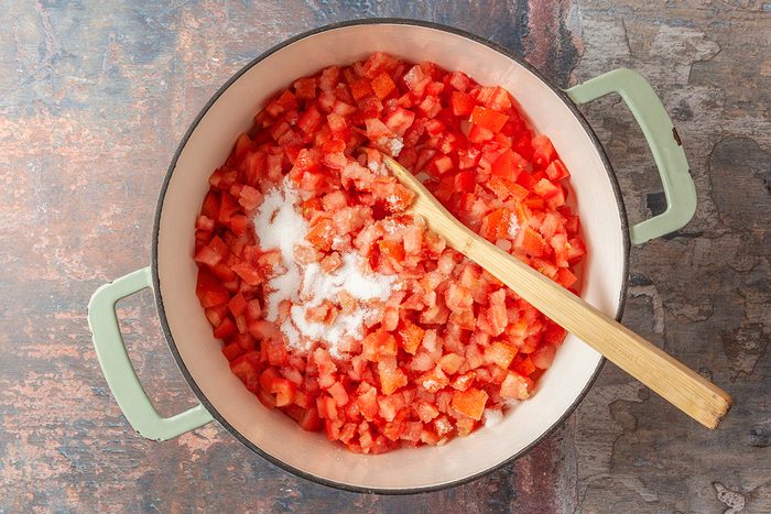 This is a top-down shot showing a large pot with light green handles containing diced tomatoes layered with salt; a wooden spoon rests inside; the pot sits on a rustic textured background