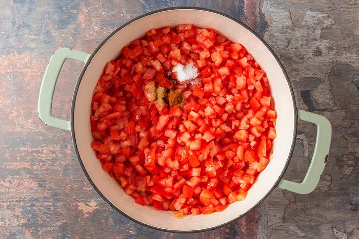 A top-down shot shows a large pot with light green handles; filled with chopped tomatoes; salt; and spices on a rustic surface; ingredients are ready to mix or cook