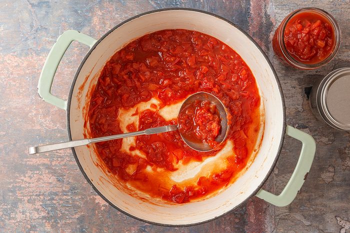 This is an overhead shot showing a white pot with light green handles full of chunky tomato sauce; a metal ladle partially removes the sauce; next to the pot are a filled jar; an empty jar; and its lid
