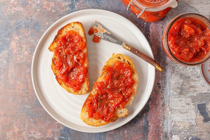 This is a close-up food shot; two slices of toasted bread with chunky tomato chutney rest on a white plate; nearby are chutney jars and a small knife on a rustic background