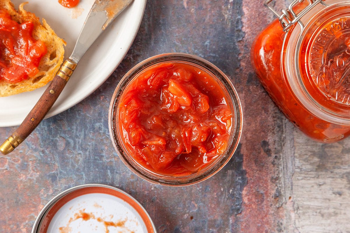 This is an overhead shot featuring a jar of chunky tomato chutney; a slice of bread with chutney on a plate; a knife; extra chutney jar; and jar lid arranged on a rustic surface