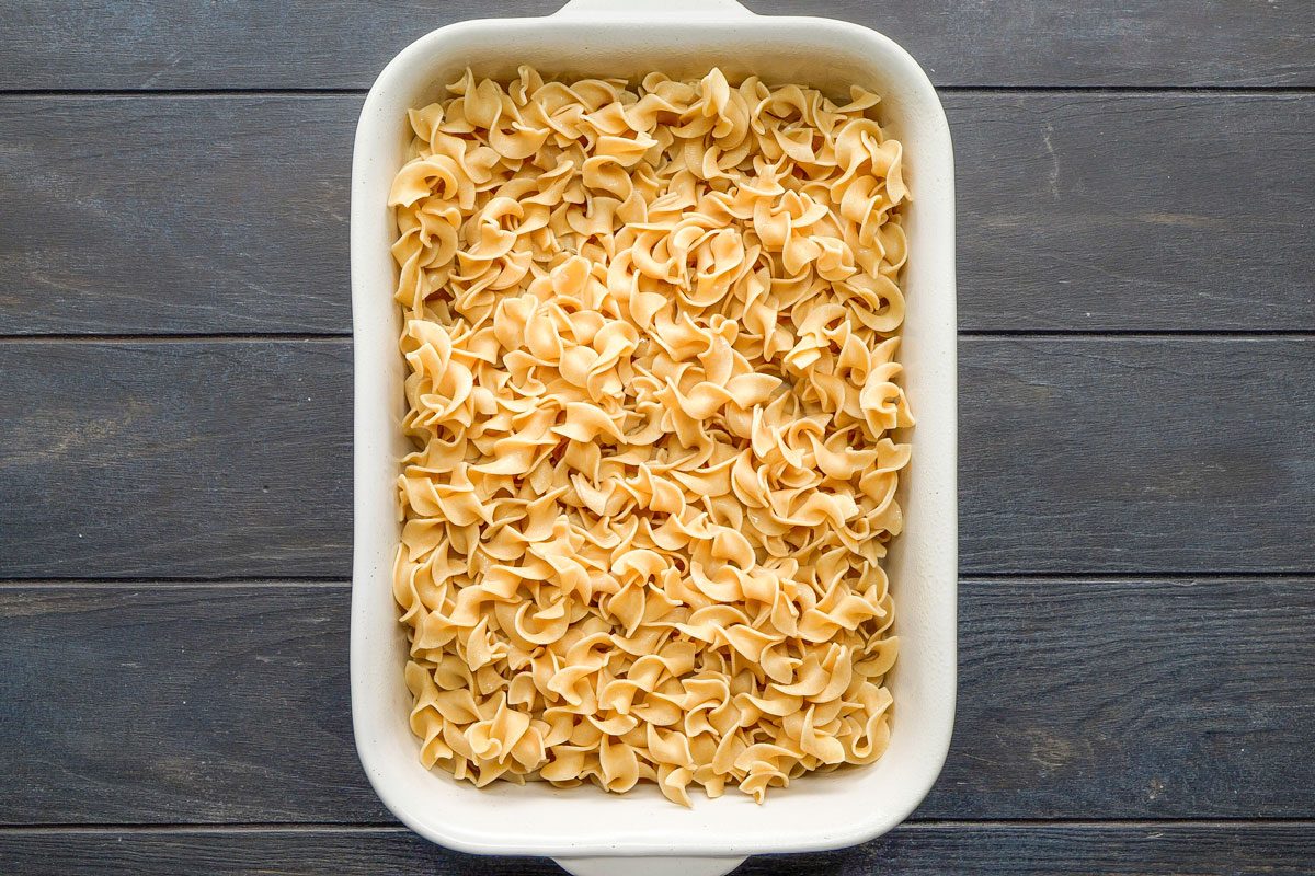A Top Shot of the image shows a white rectangular baking dish filled with uncooked egg noodles on a dark wooden surface, viewed from above; the contrast highlights the noodles texture; the scene appears ready for cooking or preparation
