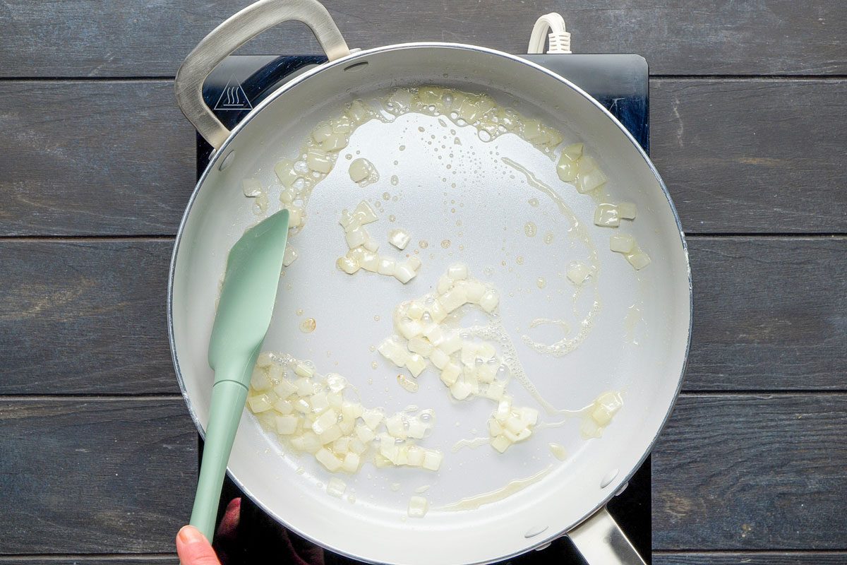 Top Shot of a hand stirs diced onions in a white frying pan with a green spatula on a stovetop; onions sauté in oil, emitting aroma