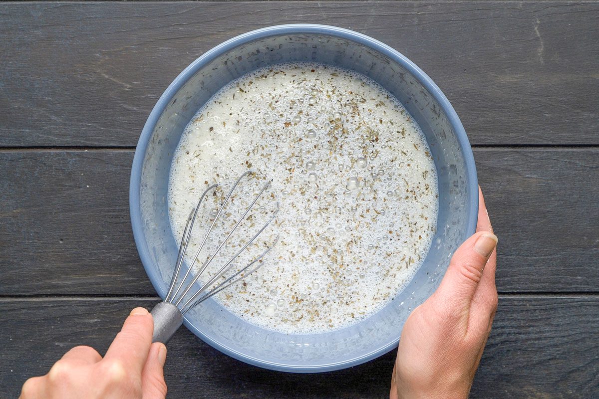 Top Shot of a person whisks a creamy; pepper-speckled mixture in a blue bowl over a dark wooden surface; showing focused food preparation and culinary action with visible textures and hand involvement