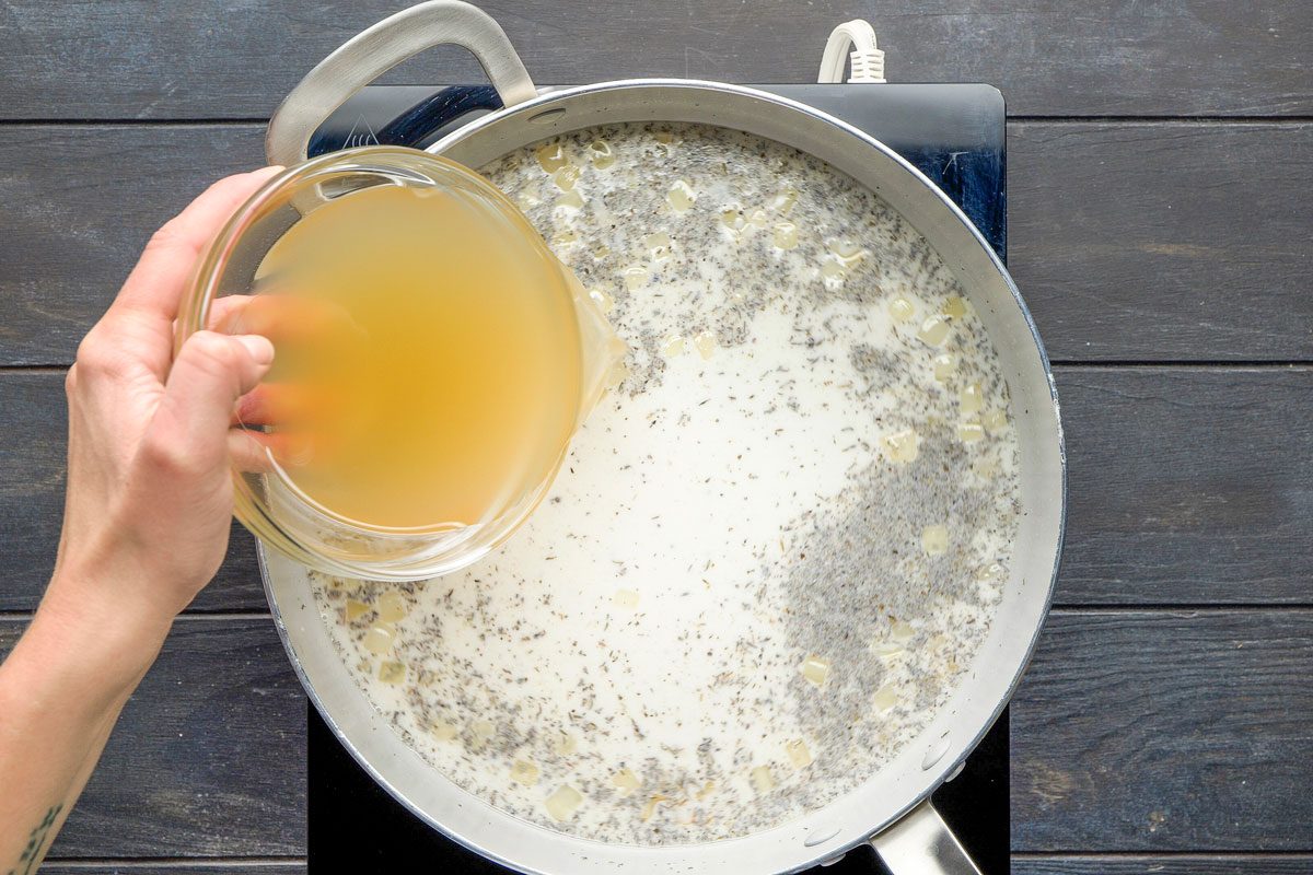 Top Shot of a hand pours broth from a glass bowl into a pan of creamy herb mixture, simmering on a stovetop placed on a wooden surface; creating a warm; home-cooked atmosphere