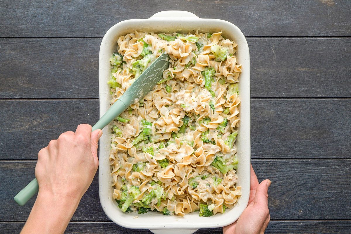 Top Shot of a person stirs creamy pasta and broccoli in a casserole with a green spatula; the dish rests on a dark wooden surface, creating a cozy cooking scene