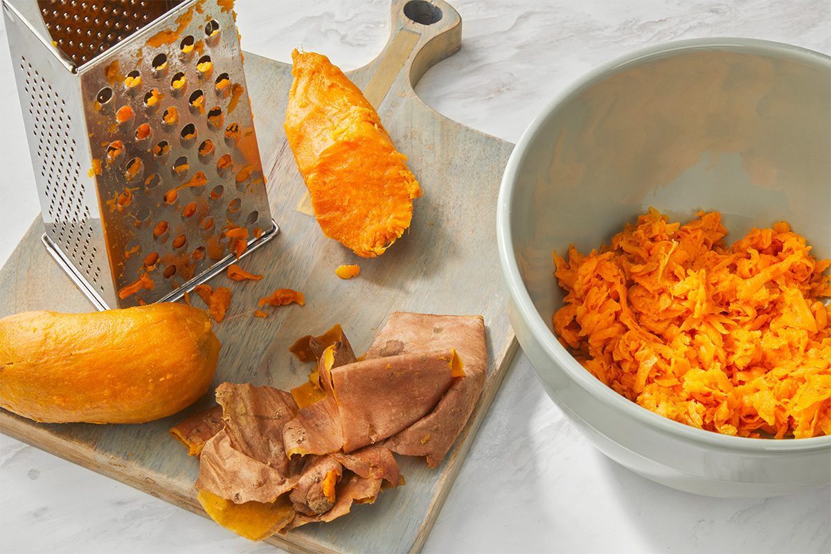 A cutting board with a metal grater, peeled and unpeeled sweet potatoes, sweet potato peels, and a bowl filled with grated sweet potato on a light surface.