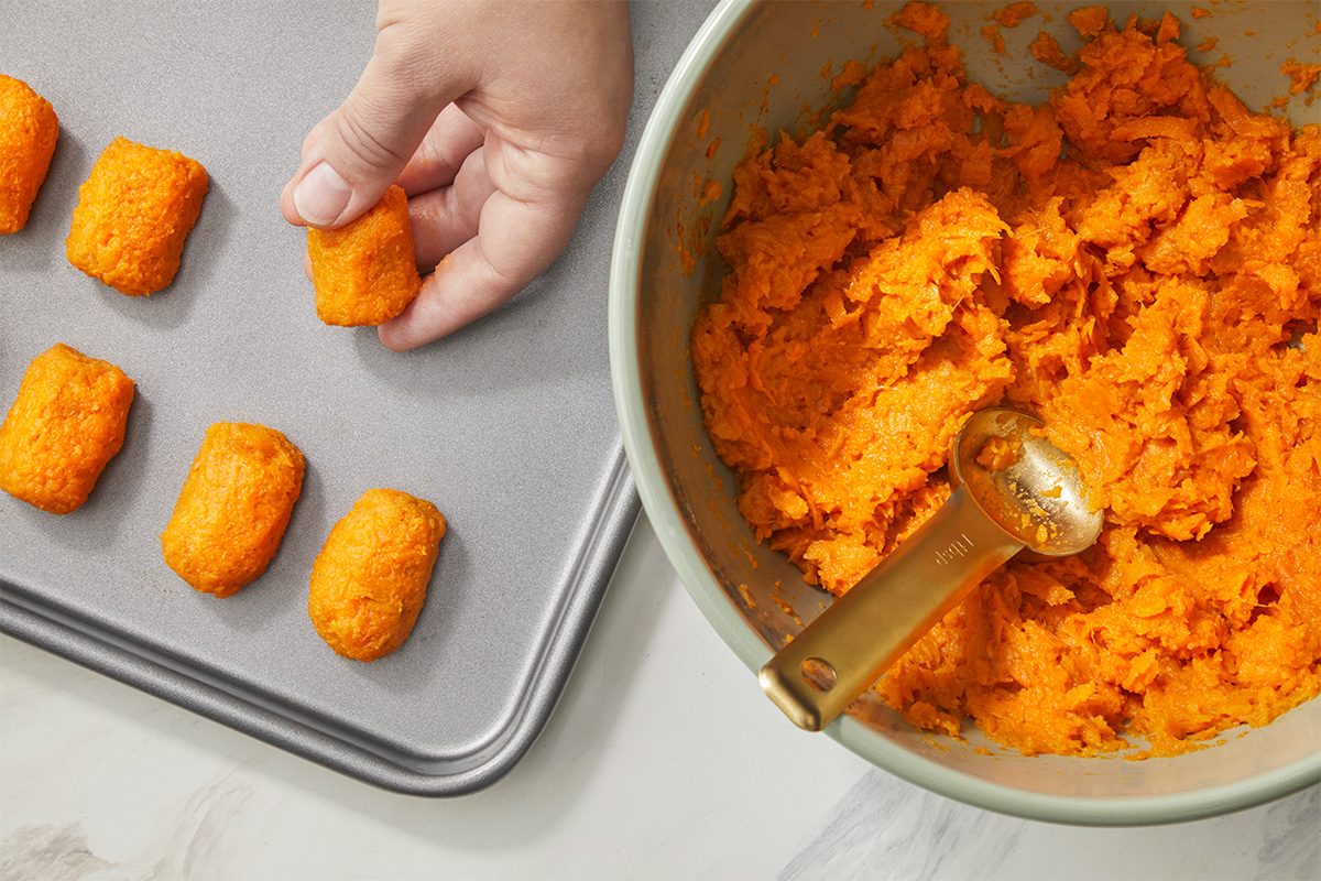 A hand places a rectangular orange nugget onto a baking sheet, next to a bowl filled with orange mashed mixture and a gold measuring spoon.