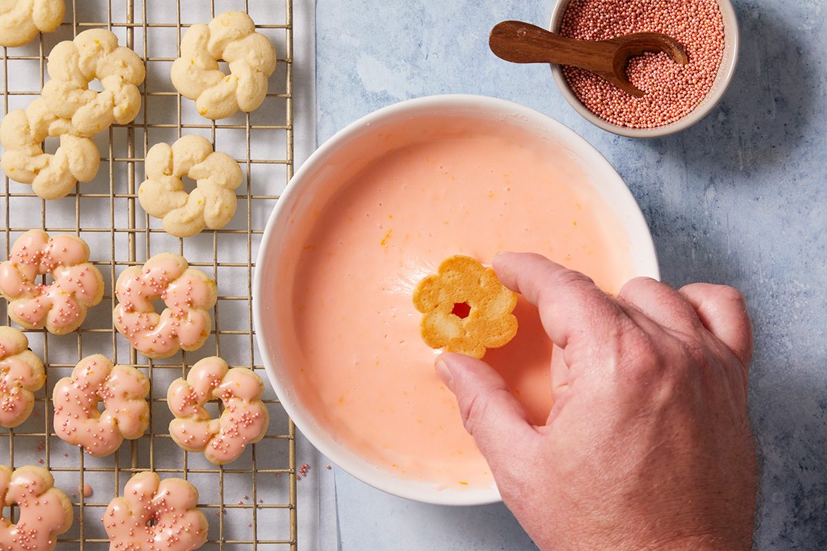 A hand dips a flower-shaped cookie into a bowl of pink icing, with more cookies on a cooling rack and a small bowl of pink sprinkles nearby.