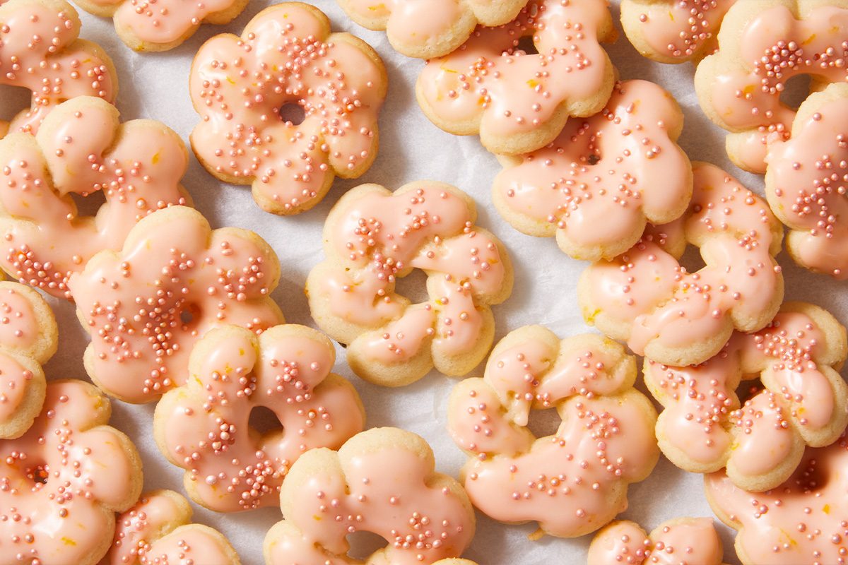 A batch of flower-shaped cookies with light pink icing and pink sprinkles, arranged closely together on a white parchment paper background.
