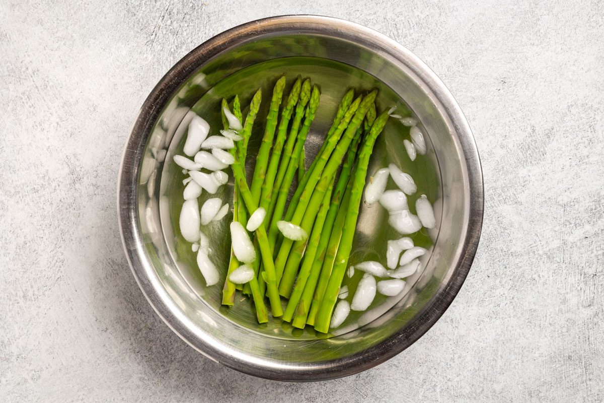 Overhead shot; a metal bowl containing blanched asparagus spears and ice cubes; on a light gray surface