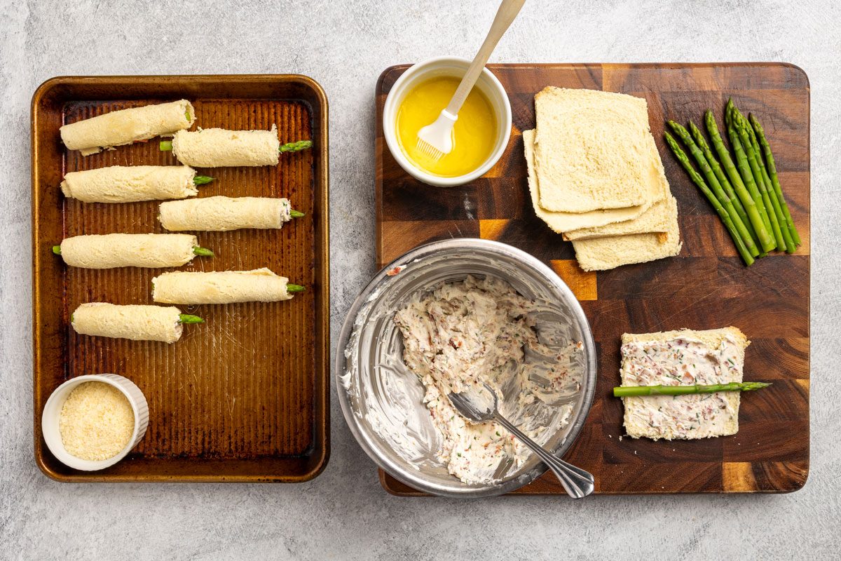 Top-down shot; a wooden board with bread slices; asparagus; bowl of spread; tray with rolled bread; melted butter; brush; grated cheese