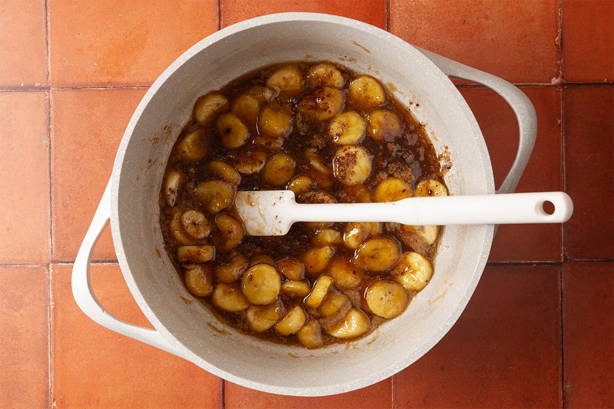 A white pot with sliced bananas cooking in a brown, syrupy sauce, stirred with a white spatula, sits on an orange tiled surface.