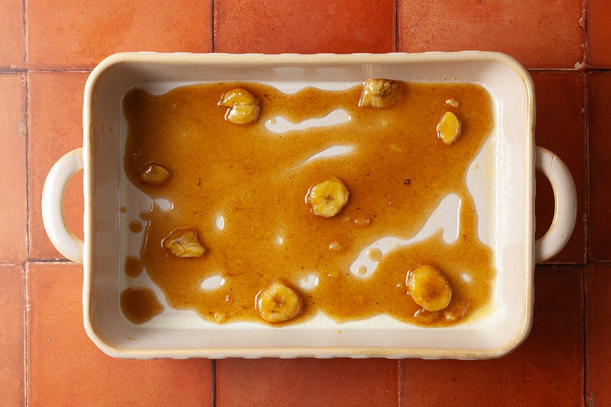 A rectangular white baking dish with handles contains a caramel-like sauce and a few sliced bananas, placed on a terracotta tiled surface.