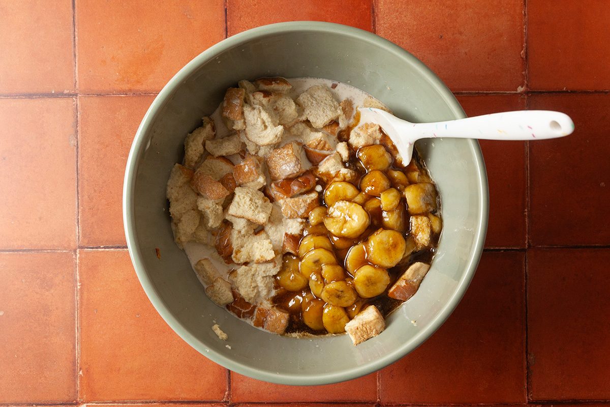 A green bowl on a red tiled surface, filled with chunks of bread, sliced bananas, and a brown syrup, with a white spoon resting inside.