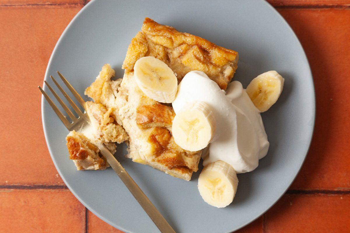 A slice of banana bread pudding topped with whipped cream and banana slices on a light blue plate, with a fork beside the dessert. The plate sits on a reddish-brown tiled surface.