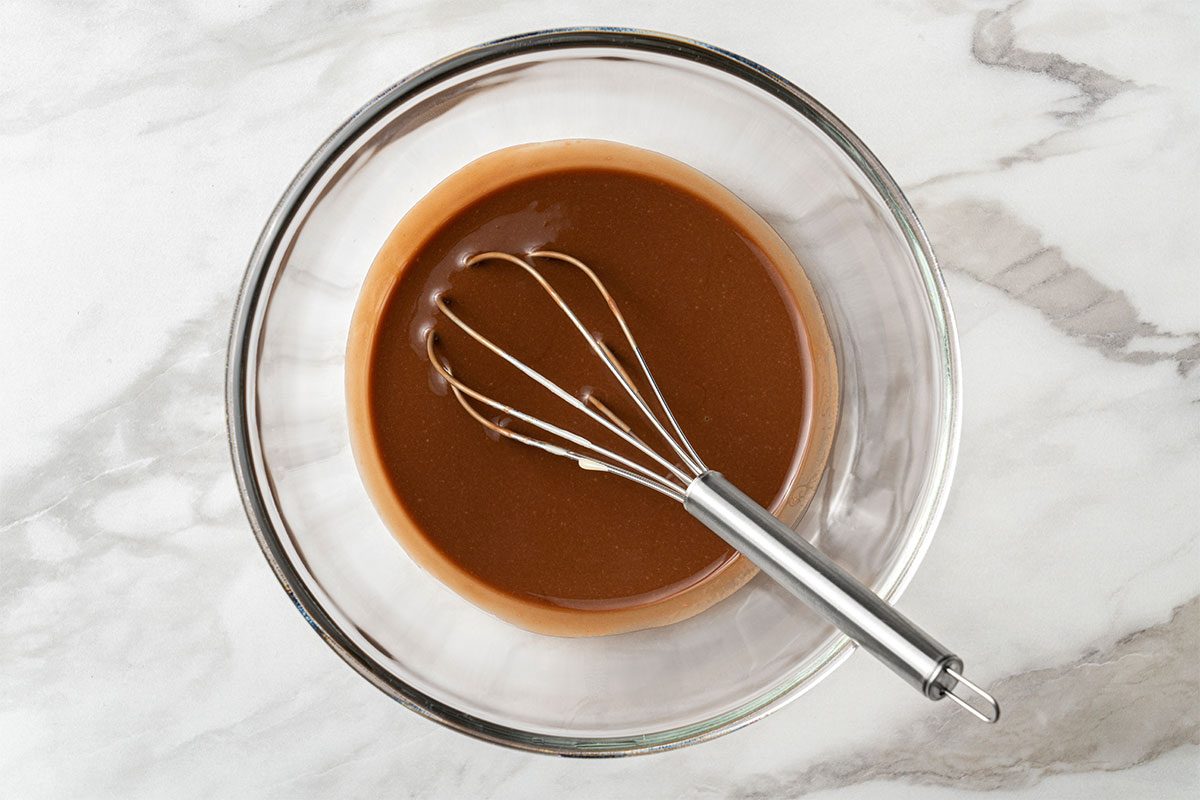 A metal whisk rests in a glass bowl filled with smooth, light brown chocolate mixture on a white marble surface.
