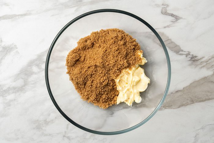 A clear glass bowl on a marble surface contains light brown sugar and a portion of soft, pale yellow butter placed side by side.