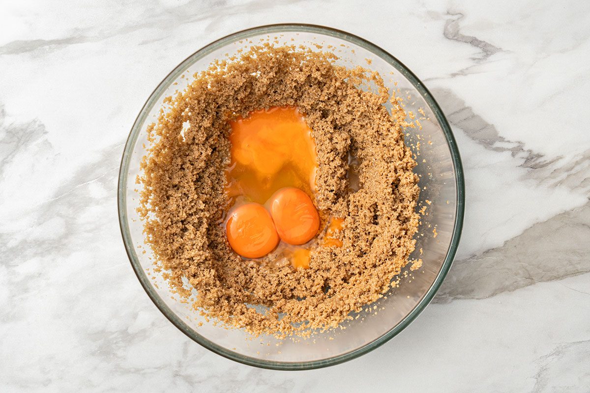 A glass bowl containing brown sugar mixture with two egg yolks and whites cracked in the center, ready to be mixed, on a marble countertop.