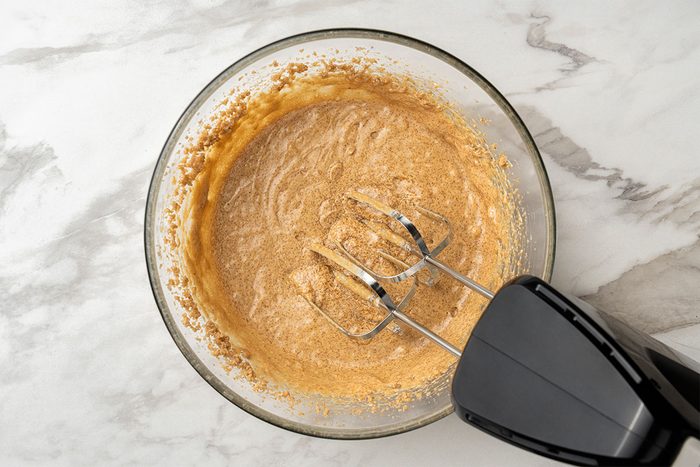 A glass bowl filled with a light brown batter is being mixed with a black electric hand mixer on a white marble countertop.