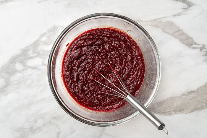 A glass bowl filled with thick, dark red sauce or puree sits on a marble surface. A metal whisk rests inside the bowl.