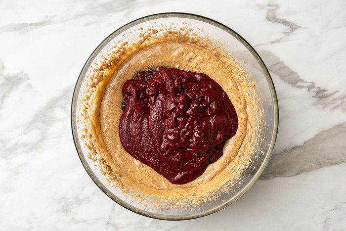 A glass bowl on a marble surface contains light brown batter topped with a thick layer of dark red fruit puree or sauce.