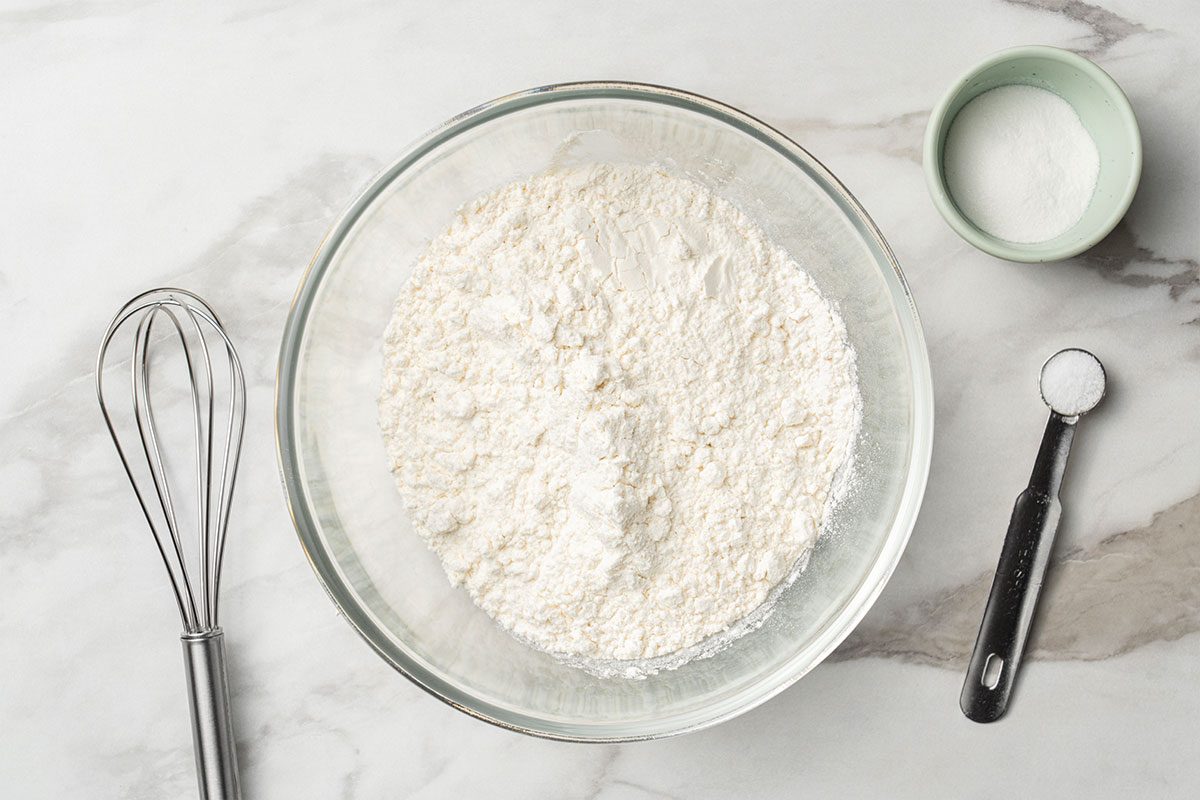 A glass bowl filled with flour sits on a marble countertop next to a whisk, a measuring spoon with baking powder, and a small bowl of sugar.