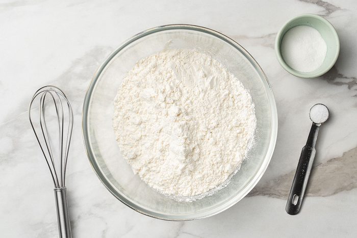 A glass bowl filled with flour sits on a marble countertop next to a whisk, a measuring spoon with baking powder, and a small bowl of sugar.