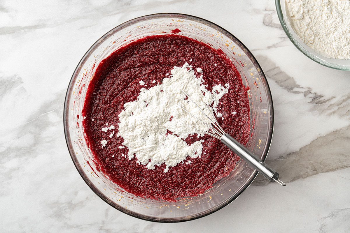 A glass bowl filled with red batter, topped with a mound of white flour, sits on a marble surface. A metal whisk rests inside the bowl, ready to mix the ingredients together.