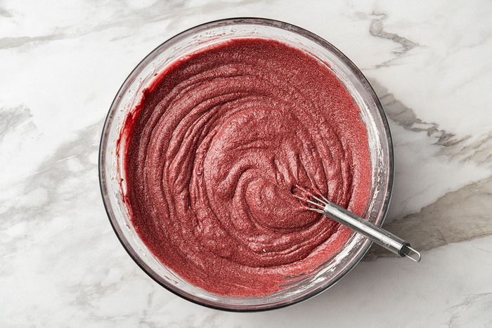 A glass bowl filled with red cake batter being mixed with a metal whisk, placed on a white marble countertop.