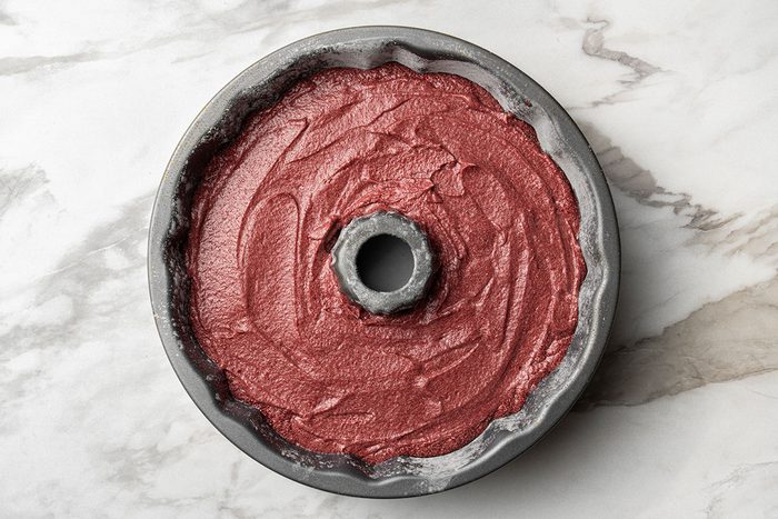 A Bundt pan filled with unbaked red cake batter sits on a white marble surface, ready to go into the oven.