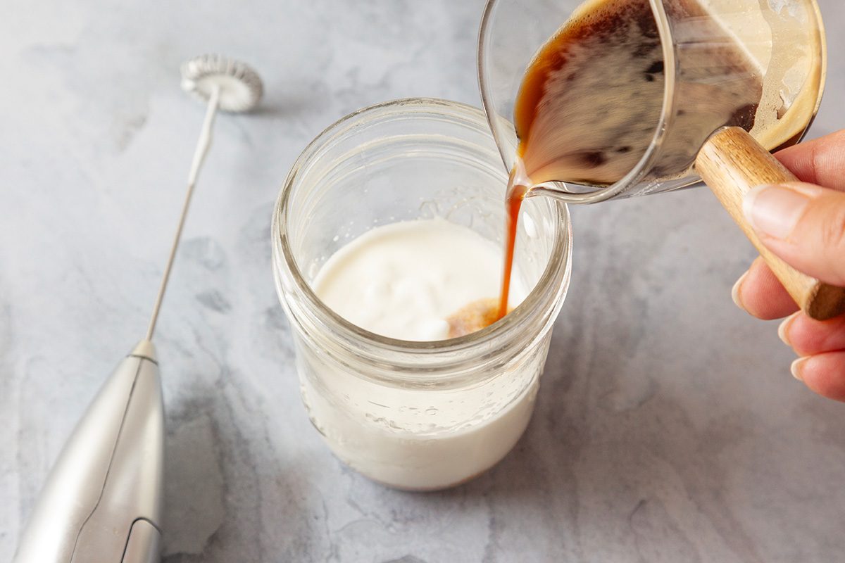 Overhead shot of a hand pours espresso into a jar of frothed milk on a light countertop; with a handheld milk frother lying nearby