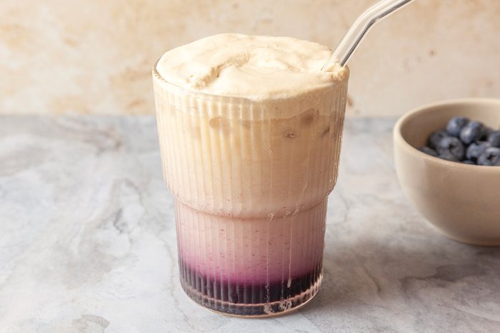 Close-up shot of a glass of Blueberry Cloud Coffee with a frothy top and a layer of purple syrup at the bottom; served with a glass straw; a bowl of blueberries is in the background