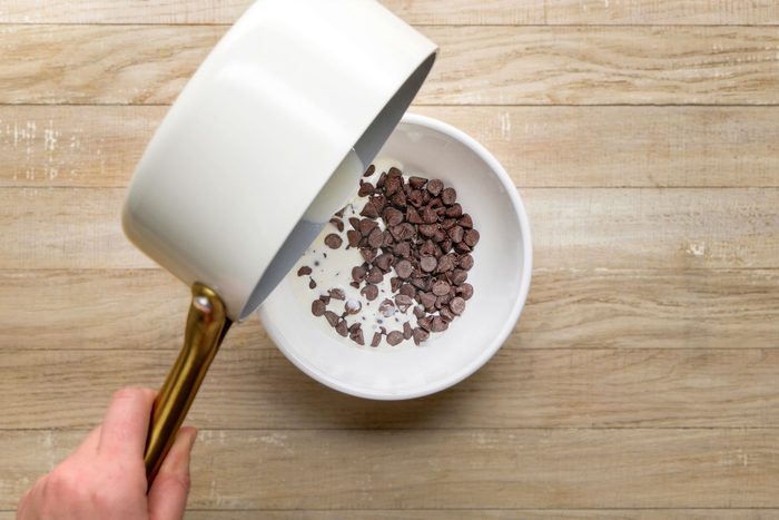 overhead shot of a hand pours liquid from a white saucepan into a white bowl filled with chocolate chips, on a light wooden surface