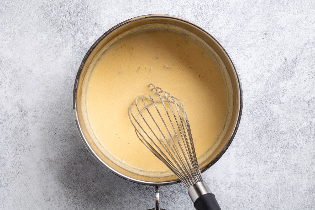 Overhead shot; metal saucepan with yellow custard and a metal whisk; set on light gray textured background