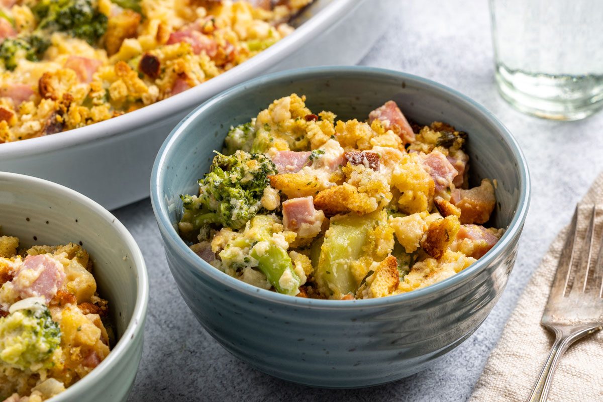 This is an overhead shot; a blue bowl with broccoli; ham; bread crumb casserole sits near a fork; napkin; extra serving dish