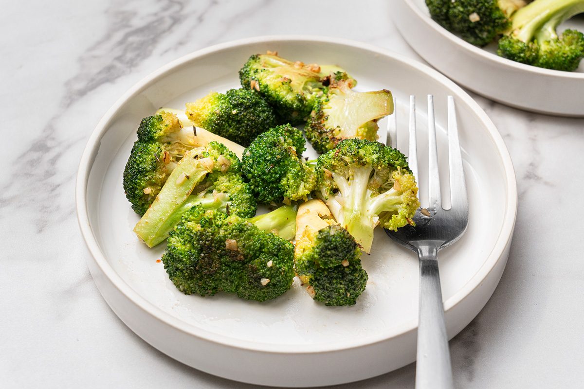 A white plate with roasted broccoli florets, lightly seasoned, next to a fork. The plate is set on a white marble surface, with a second plate partially visible in the background.
