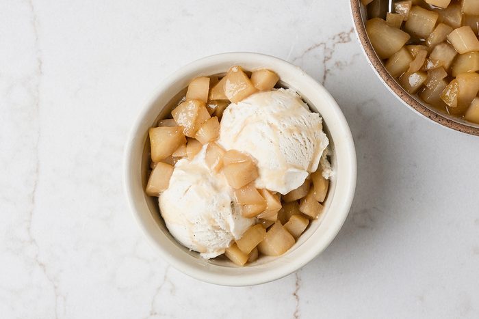 A bowl with two scoops of vanilla ice cream topped with small diced caramelized apples, photographed on a white marble surface. Another bowl with diced apples is partially visible in the corner.