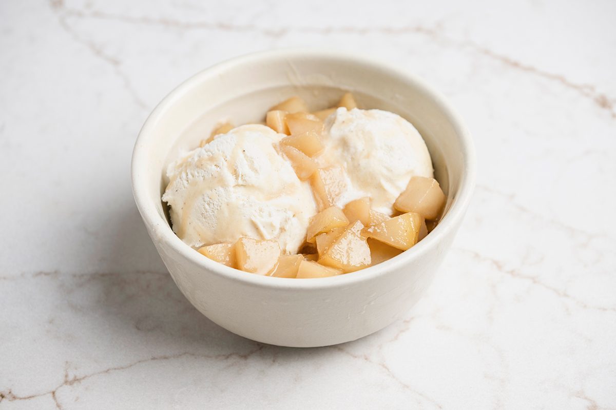 A white bowl containing two scoops of vanilla ice cream topped with small pieces of cooked apples, set on a white marble surface.