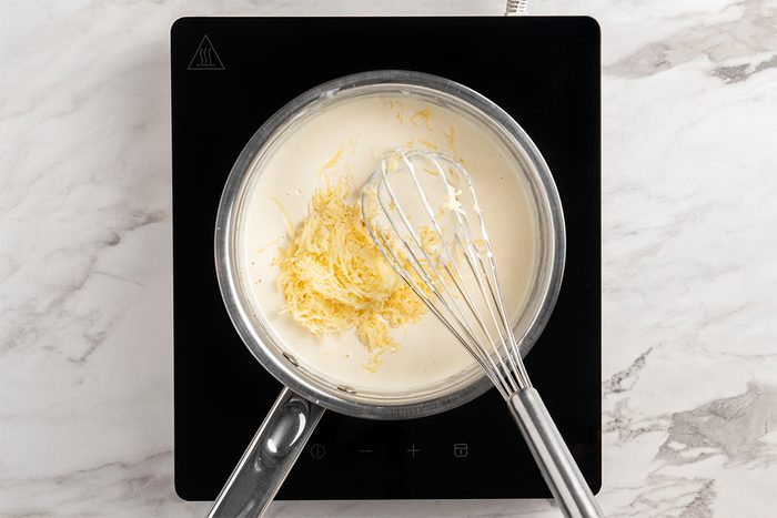 A saucepan with creamy white sauce and shredded cheese being whisked on a black induction cooktop, set on a light marble countertop.