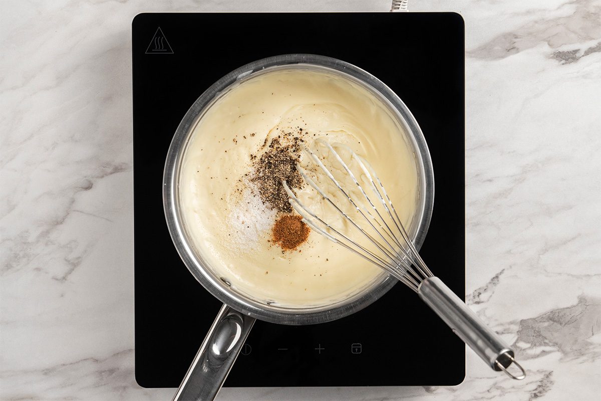 A saucepan with creamy white sauce is on a stovetop. A whisk is stirring in black pepper and spices on the sauce’s surface. The scene is viewed from above on a marble countertop.