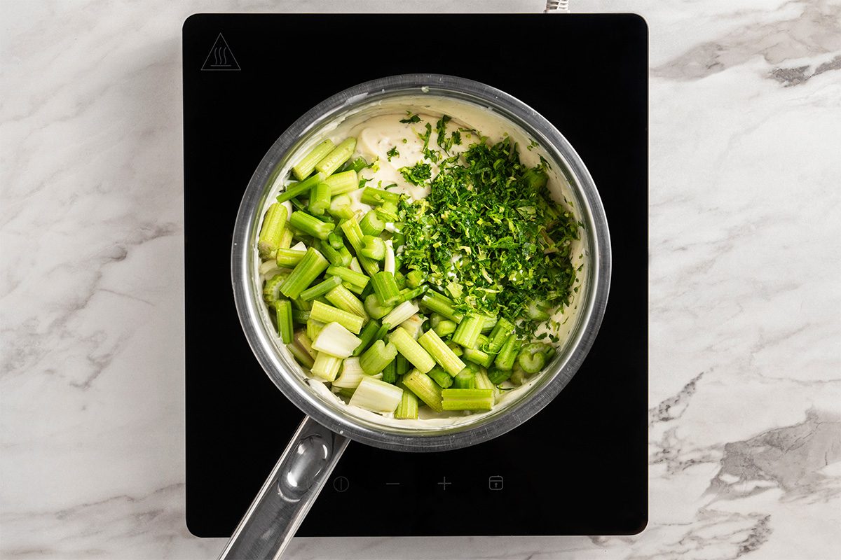 A saucepan on an induction cooktop contains chopped green onions and fresh herbs on top of a creamy mixture, ready to be stirred. The scene is set on a white marble countertop.