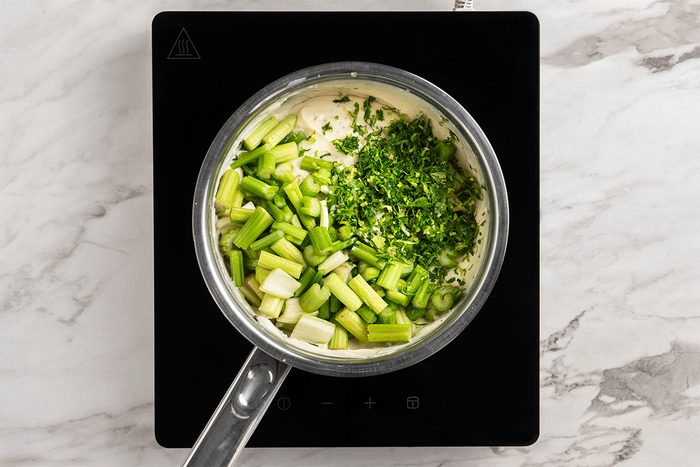 A saucepan on an induction cooktop contains chopped green onions and fresh herbs on top of a creamy mixture, ready to be stirred. The scene is set on a white marble countertop.
