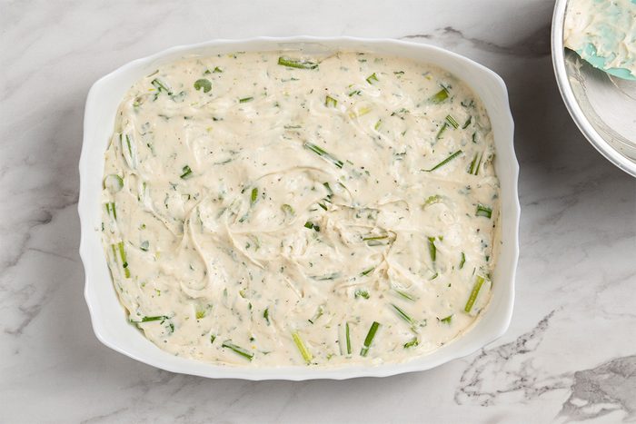 A white baking dish filled with an uncooked creamy mixture containing chopped green herbs and vegetables, resting on a light marble surface.