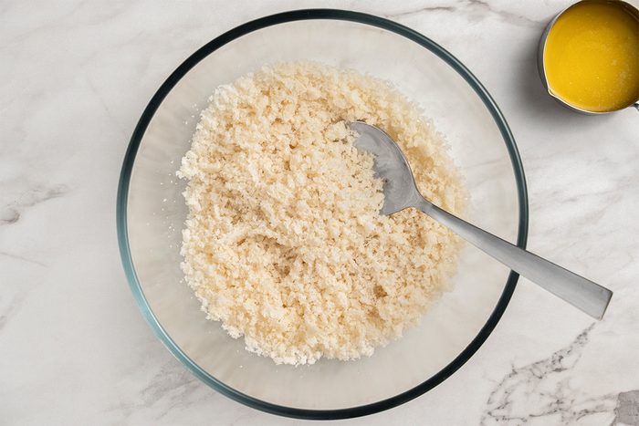 A glass bowl filled with breadcrumbs and a spoon on a marble surface; a small metal cup with melted butter sits nearby.