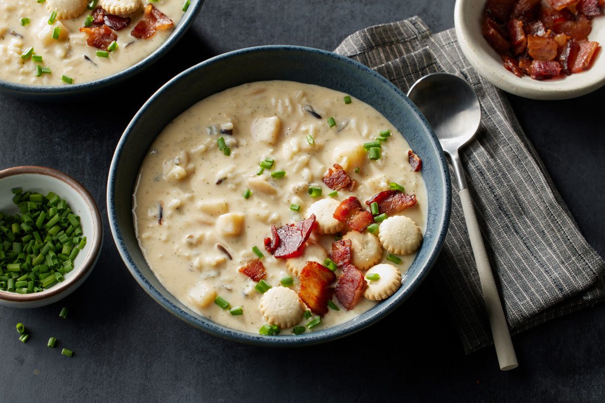 overhead shot of a bowl of creamy chowder topped with bacon pieces, oyster crackers, and chopped chives sits on a table next to a spoon, a bowl of bacon, and a small dish of chives